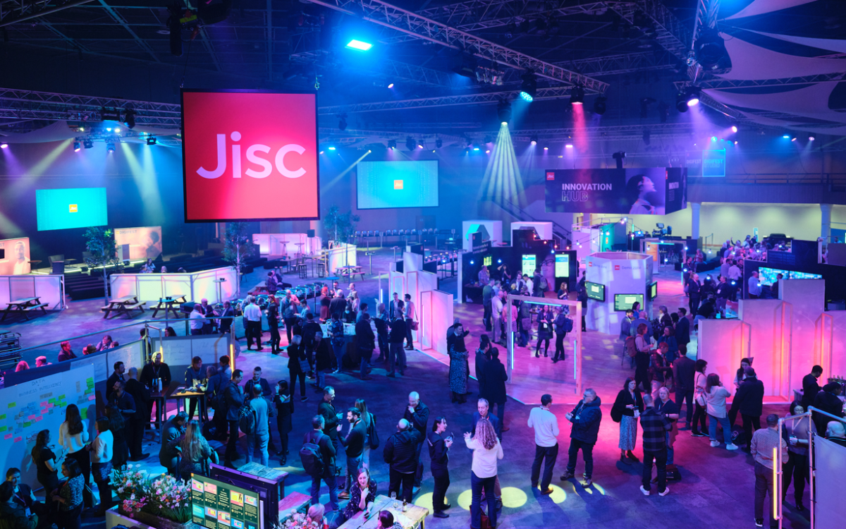 A large Jisc sign hangs above a busy auditorium where people are talking and looking at stall areas. Mood lighting illuminates the hall