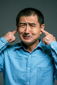 A man in a blue shirt with fingers in his ears because of noise.